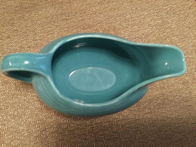 May include: A turquoise blue ceramic gravy boat with a smooth, rounded interior and a slightly flared lip. The boat has a single handle on the left side.