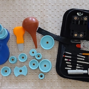 May include: A blue metal watch press tool with a long handle, a brown bulb blower, various light blue circular dies, and a black zippered case containing assorted watch repair tools, including screwdrivers, tweezers, and a metal case opener.