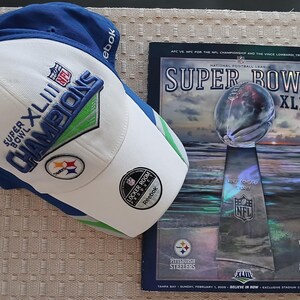 Könnte beinhalten: Eine blau-weiße Reebok Baseballkappe mit Super Bowl XLIII Champions Stickerei und Pittsburgh Steelers Logo, neben einem Super Bowl XLIII Programmheft, das die Vince Lombardi Trophäe und Teamlogos auf einem dunklen Hintergrund zeigt.