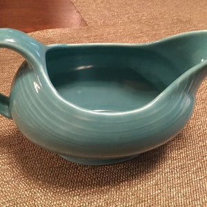 May include: A turquoise blue ceramic gravy boat with a wide, rounded base and a handle. The boat has a smooth, glossy finish.