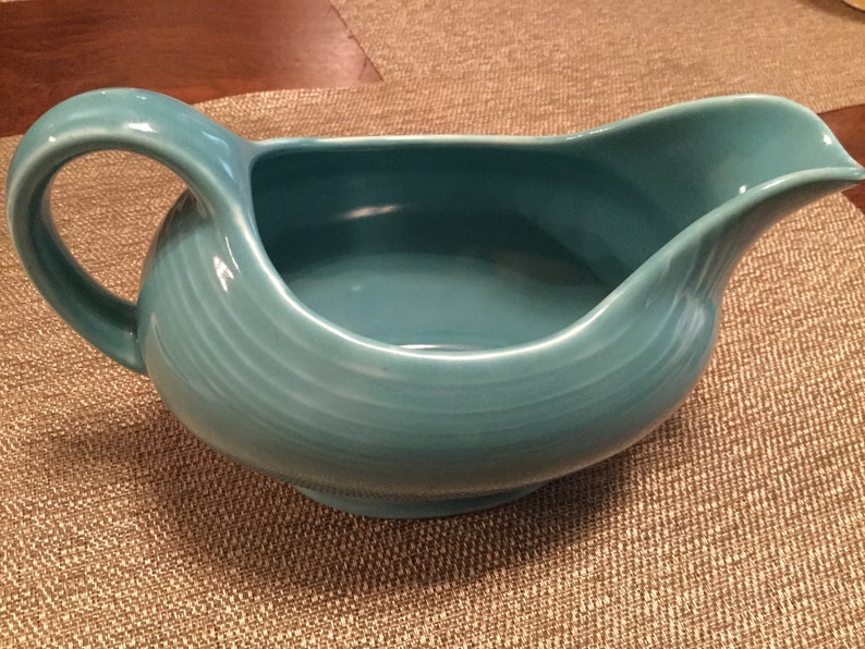 May include: A turquoise blue ceramic gravy boat with a wide, rounded base and a handle. The boat has a smooth, glossy finish.