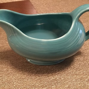May include: A turquoise ceramic gravy boat with a handle. The boat has a smooth, rounded shape and a slightly flared lip.