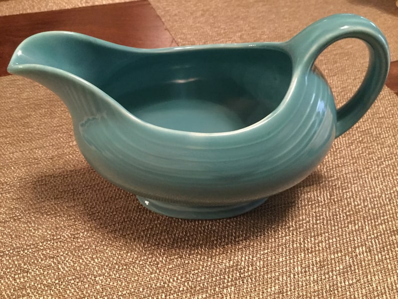 May include: A turquoise ceramic gravy boat with a handle. The boat has a smooth, rounded shape and a slightly flared lip.