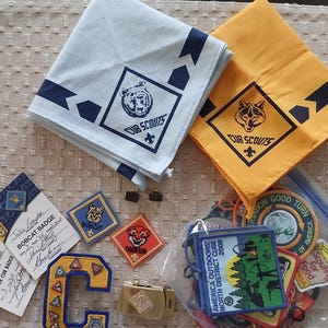 May include: A collection of Cub Scouts memorabilia including a light blue neckerchief with a bear logo, a yellow neckerchief with a wolf logo, various embroidered patches, two signed Bobcat and Tiger Cub badge cards, and a gold-tone belt buckle.