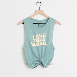 May include: A sleeveless, teal-colored tank top with the text "I RUN THIS 'HOOD." in a retro font. The bottom of the tank top is knotted. The tank top is hanging on a wooden hanger against a white background.