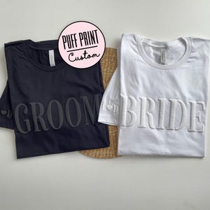 May include: Two t-shirts with puff print text. One black t-shirt says "Groom" and one white t-shirt says "Bride".
