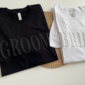 May include: Two t-shirts, one black and one white, with the words "Groom" and "Bride" in raised lettering.