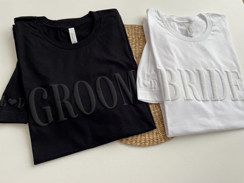 May include: Two t-shirts, one black and one white, with the words "Groom" and "Bride" in raised lettering.