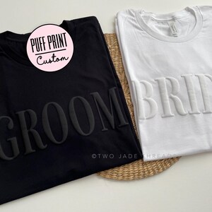 May include: Two black and white t-shirts with the words "Groom" and "Bride" in raised lettering. The t-shirts are made of a soft, comfortable material and are perfect for a wedding or engagement party.