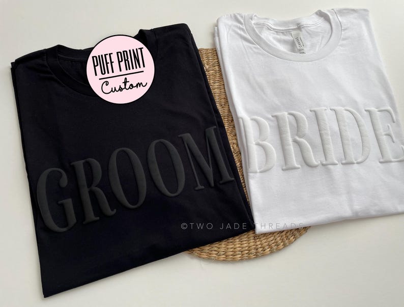 May include: Two black and white t-shirts with the words "Groom" and "Bride" in raised lettering. The t-shirts are made of a soft, comfortable material and are perfect for a wedding or engagement party.