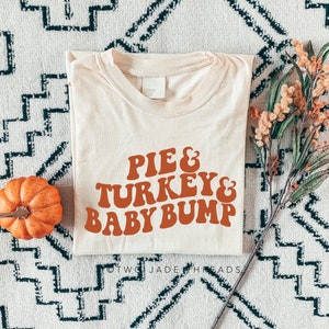 May include: Cream-colored t-shirt with the text "PIE & TURKEY & BABY BUMP" in orange retro font. A small orange pumpkin and fall foliage are placed near the shirt on a patterned rug. The shirt is folded.