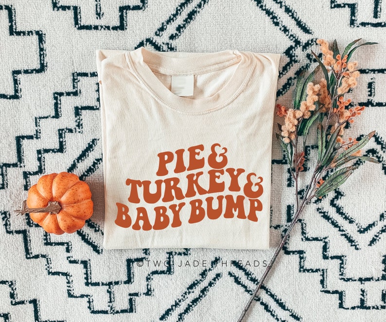 May include: Cream-colored t-shirt with the text "PIE & TURKEY & BABY BUMP" in orange retro font. A small orange pumpkin and fall foliage are placed near the shirt on a patterned rug. The shirt is folded.
