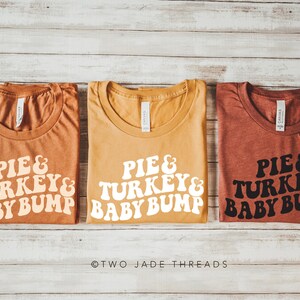 May include: Three t-shirts in shades of orange, yellow, and red-brown, each with the text "PIE & TURKEY & BABY BUMP" in a retro font. The shirts are laid flat on a light wood-grain surface.