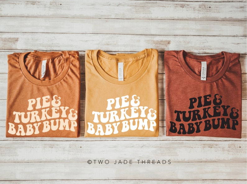 May include: Three t-shirts in shades of orange, yellow, and red-brown, each with the text "PIE & TURKEY & BABY BUMP" in a retro font. The shirts are laid flat on a light wood-grain surface.