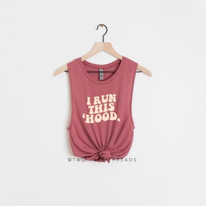 May include: A dusty rose muscle tank top with the text "I RUN THIS 'HOOD" in a retro font. The tank top is knotted at the waist and hangs on a wooden hanger against a white background.