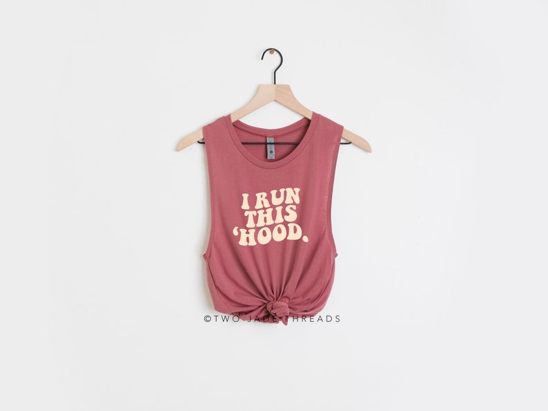May include: A dusty rose muscle tank top with the text "I RUN THIS 'HOOD" in a retro font. The tank top is knotted at the waist and hangs on a wooden hanger against a white background.