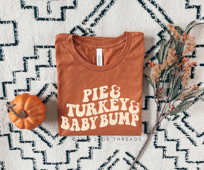 May include: An orange-brown t-shirt with the words "PIE & TURKEY & BABY BUMP" in a retro font. A small pumpkin and decorative foliage are placed near the shirt on a patterned rug. The shirt is from Two Jade Threads.