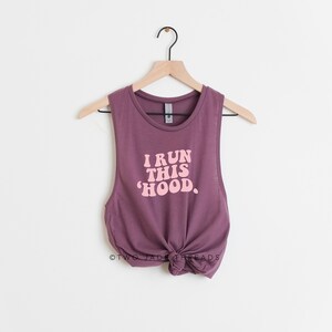 May include: A mauve-colored tank top with the text "I RUN THIS 'HOOD." in a retro font. The sleeveless top is knotted at the bottom and hangs on a wooden hanger against a white background. The tank top is a casual wear item.