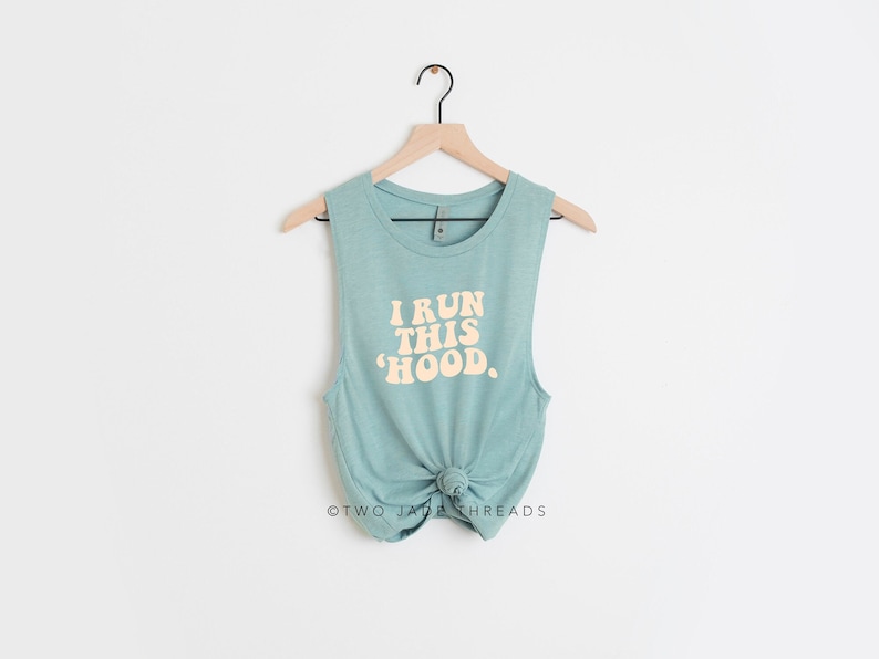 May include: A sleeveless, teal-colored tank top with the text "I RUN THIS 'HOOD." in a retro font. The bottom of the tank top is knotted. The tank top is hanging on a wooden hanger against a white background.