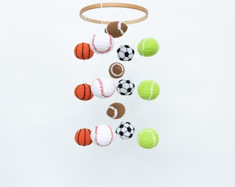 May include: A white wooden mobile with crocheted sports balls hanging from it. The balls are a baseball, basketball, football, soccer ball, and tennis ball.
