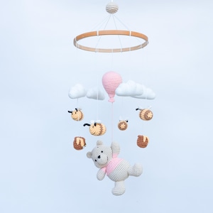 Könnte beinhalten: Ein gehäkeltes Baby-Mobile mit einem rosa Ballon, weißen Wolken und braunen und gelben Bienen. Ein gehäkelter Teddybär hängt am unteren Ende des Mobiles.