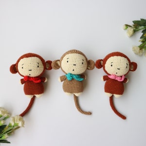 May include: Three crocheted monkey toys with brown fur, white faces, and different colored scarves. The monkeys are sitting on a white background.