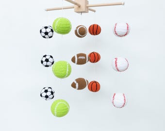 May include: A wooden baby mobile with crocheted sports balls, including a baseball, basketball, football, soccer ball, and tennis ball. The balls are suspended from a wooden frame.