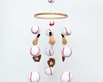 May include: A white and red crocheted baseball themed baby mobile with a wooden ring. The mobile features baseballs, bats, and a glove.