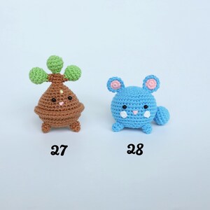 May include: Two crocheted plush toys. One is brown with green accents and the number 27 below it. The other is blue with pink accents and the number 28 below it. Both have facial features.