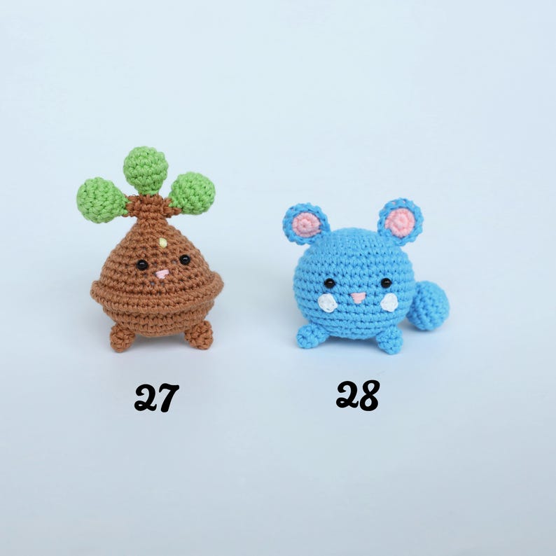 May include: Two crocheted plush toys. One is brown with green accents and the number 27 below it. The other is blue with pink accents and the number 28 below it. Both have facial features.