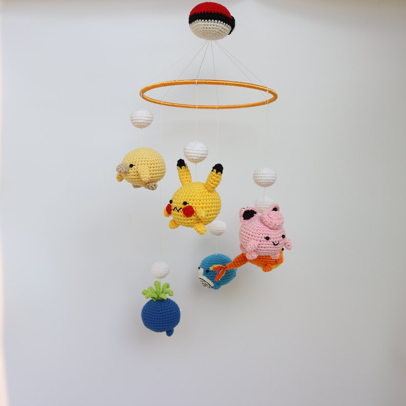 May include: A handmade baby mobile featuring crocheted Pok&eacute;mon characters. The mobile includes a yellow Pikachu, a pink Jigglypuff, a blue and orange fish, and other colorful crocheted elements. The mobile is suspended from a yellow ring and a red and white ball.