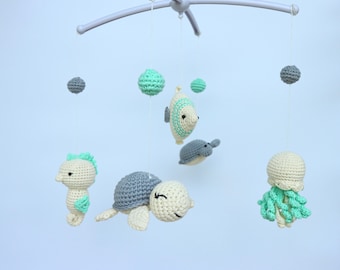 May include: A handmade baby mobile featuring crocheted sea creatures, including a seahorse, turtle, fish, dolphin, and jellyfish. The mobile has a gray frame and includes gray and mint green accents. The sea creatures are suspended by strings.
