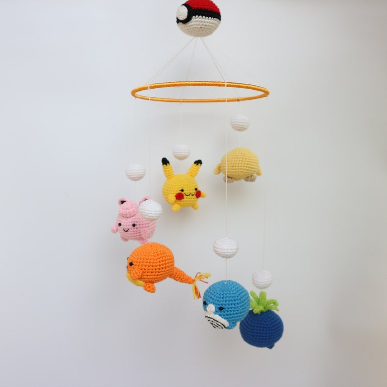 May include: A handmade crochet baby mobile featuring various colorful Pok&eacute;mon characters and white spheres suspended from a yellow ring. The mobile includes a Pok&eacute; Ball at the top and several Pok&eacute;mon characters in yellow, orange, pink, and blue.