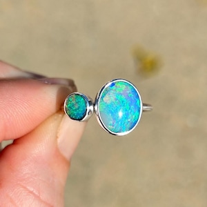 May include: A silver ring featuring two round bezel-set opals of different sizes. The larger opal displays vibrant flashes of blue and green, while the smaller adjacent opal shows a deeper, darker green tone with subtle iridescent sparkles.