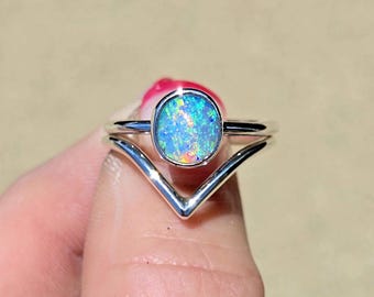 May include: A silver ring featuring a round, bezel-set opal gemstone with vibrant blue, green, and orange flashes. The band has a distinct V-shaped chevron design that frames the central stone, creating a layered appearance with a polished finish.