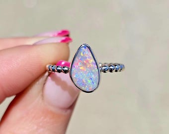 May include: A silver ring featuring a teardrop-shaped opal gemstone with iridescent flashes of pink, blue, and green. The band of the ring has a textured, beaded design that encircles the finger, providing a distinct contrast to the smooth stone.