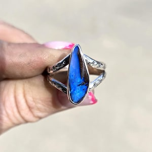 May include: A silver ring featuring a central, elongated, irregular-shaped blue and black boulder opal stone. The silver band is split into two arms, each decorated with small, stamped sunburst or floral patterns along the outer edges.