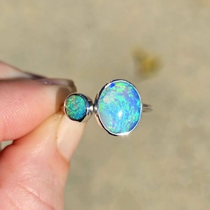 May include: A silver ring featuring two oval-shaped opals. The larger opal is a vibrant blue with green and white flashes, while the smaller one is a similar color. The ring is held up against a blurred background.