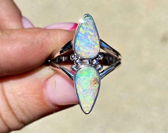 May include: A silver ring featuring two pear-shaped opal gemstones stacked vertically. The opals display a play of color with flashes of green, blue, and yellow. The metal band splits into two sections that meet at the center, accented by small silver beads.