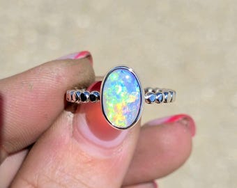 May include: A silver ring featuring an oval-shaped opal gemstone with a vibrant play of color, including flashes of green, yellow, and blue. The band of the ring has a textured, beaded design that encircles the finger, providing a distinct contrast.