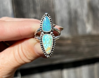 May include: A silver ring featuring two stacked, pear-shaped stones. The top stone is a vibrant blue opal with iridescent flecks, while the bottom stone is a light blue and tan turquoise with a matrix pattern. Both stones are set in beaded silver.