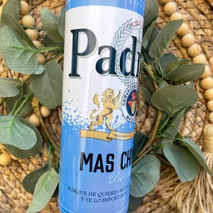 May include: A blue and white insulated tumbler with a graphic of a beer can featuring the text "Padr". The can has a lion logo and the text "Cerveceria Mexicana" and "1932". The tumbler also has the text "Mas Chido" and "Porque me quieres mucho y se lo importante".