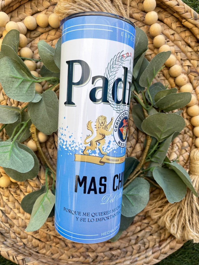 May include: A blue and white insulated tumbler with a graphic of a beer can featuring the text "Padr". The can has a lion logo and the text "Cerveceria Mexicana" and "1932". The tumbler also has the text "Mas Chido" and "Porque me quieres mucho y se lo importante".