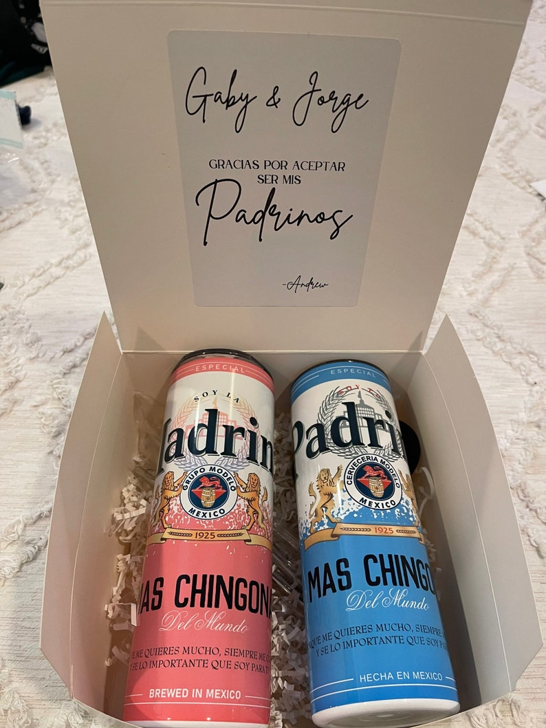 May include: Two pink and blue cans with the words "Soy la Padrino" and "Mas Chingon Del Mundo" on them. The cans are in a white box with a white card that says "Gaby & Jorge Gracias por aceptar ser mis Padrinos -Andrew".
