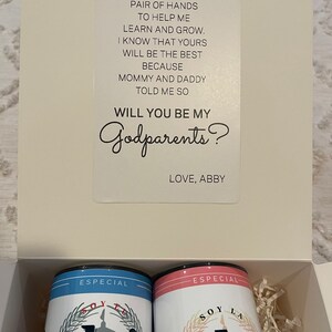 May include: A white box with a card inside that says "Will you be my Godparents?" with two blue and pink cans that say "Especial Soy La" with a building graphic. The cans are in a shredded paper box.