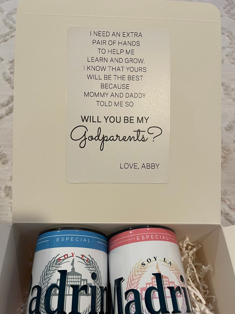 May include: A white box with a card inside that says "Will you be my Godparents?" with two blue and pink cans that say "Especial Soy La" with a building graphic. The cans are in a shredded paper box.
