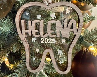 May include: A wooden Christmas ornament in the shape of a tooth, with the name "Helena" and the year "2025" cut out. The ornament has a clear center with small tooth shapes. It hangs from a natural twine string, displayed on a Christmas tree.