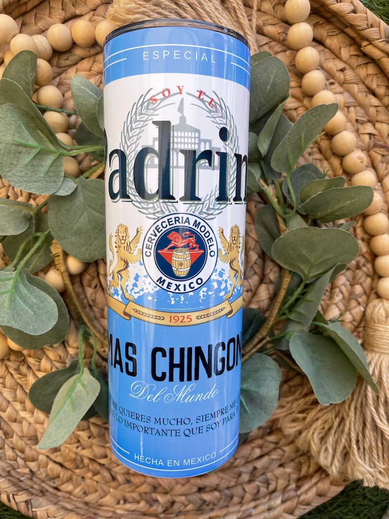 May include: A blue and white insulated tumbler with a graphic of a beer can. The can features the text "Soy El Padrino" and "Cerveceria Modelo" with a lion logo. The text "Mas Chingon Del Mundo" is printed below the can graphic.