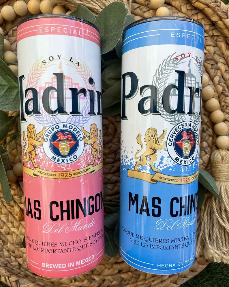 May include: Two stainless steel tumblers with a pink and blue design. The tumblers feature a beer can design with the words "Soy La Ladri" and "Soy El Padri" on the front. The tumblers also feature the words "Mas Chingon Del Mundo" and "Mas Chingon Del Mundo" on the bottom.