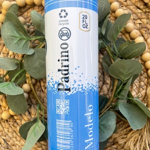 May include: A blue and white 20 fl oz tumbler with a black and white image of a beer bottle and the text "Modelo" and "Padrino". The tumbler has a recycle symbol and a no car symbol. It also has a barcode and a QR code.